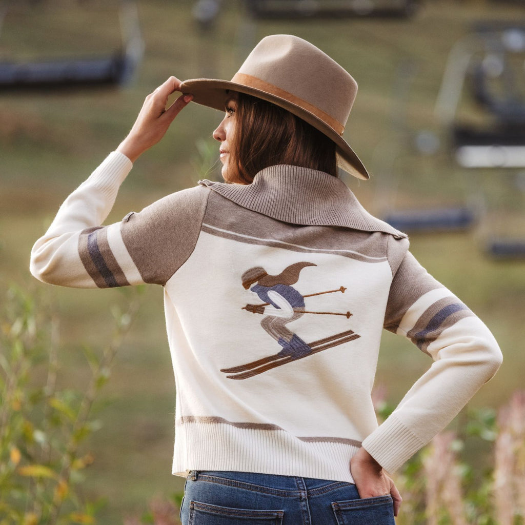 A woman wearing a cream and taupe knit sweater vest with a ski graphic on the back, paired with a brown wide-brim hat and blue jeans, photographed outdoors on a ski resort slope.