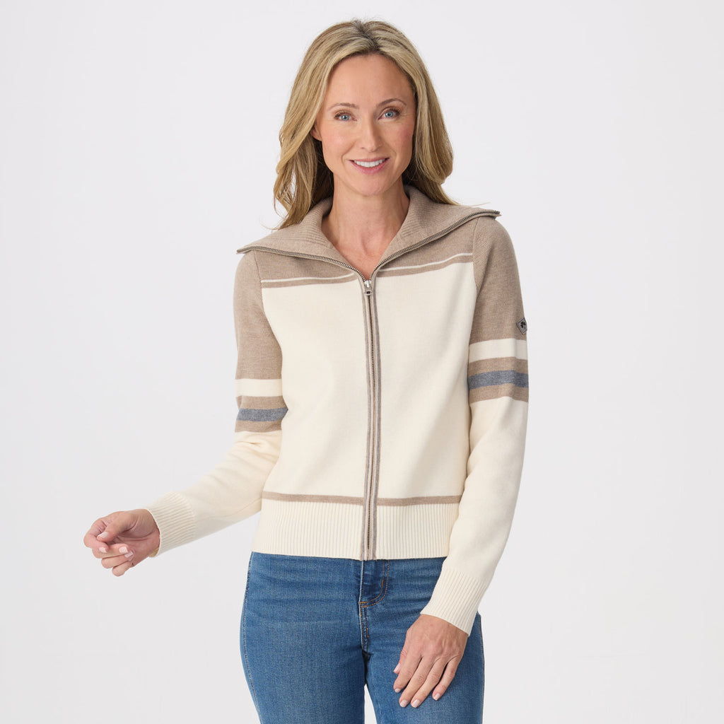 A woman modeling a cream and beige zip-up knit sweater with horizontal stripes on the chest and sleeves, paired with blue jeans, against a plain light gray studio background.
