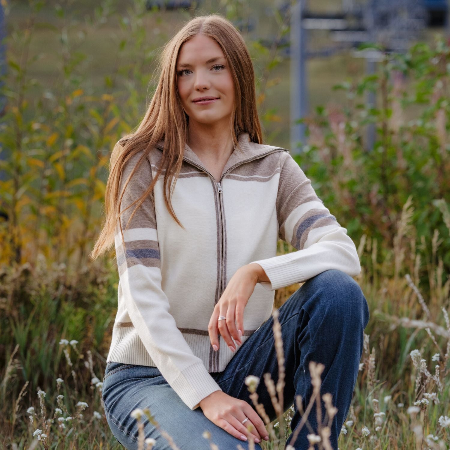 A woman with long light brown hair sits in a grassy field wearing a cream and taupe quarter-zip knit sweater with striped sleeves, paired with blue jeans.