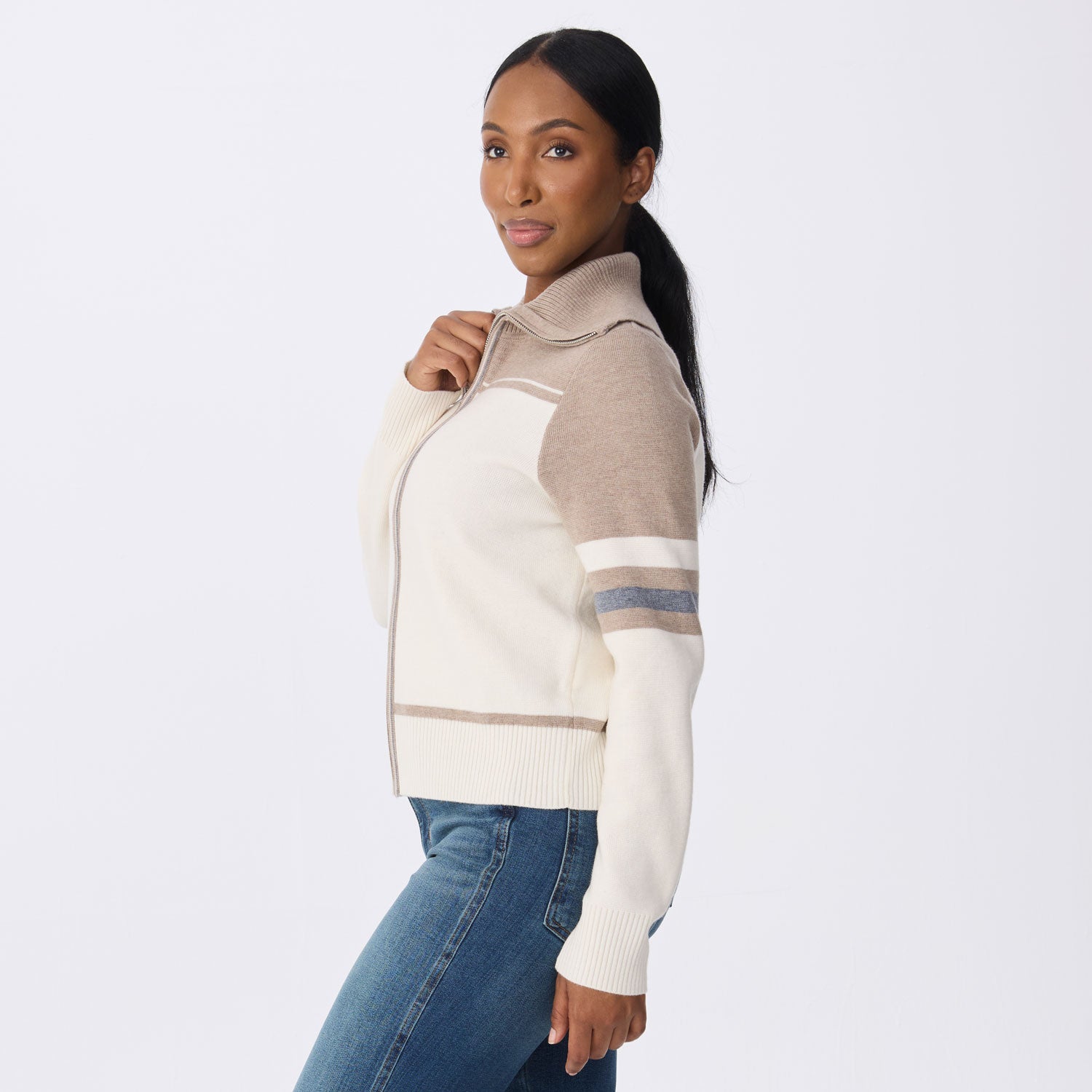 A woman modeling a cream and beige color-blocked zip-up knit sweater with blue jeans, photographed against a plain light gray studio background.