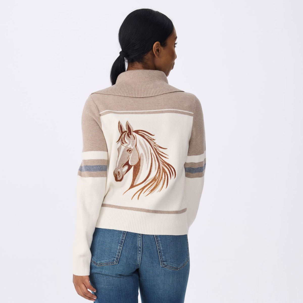 A woman shown from the back wearing a cream and beige knit sweater with a large embroidered horse head design on the back, paired with blue jeans, against a light gray studio background.