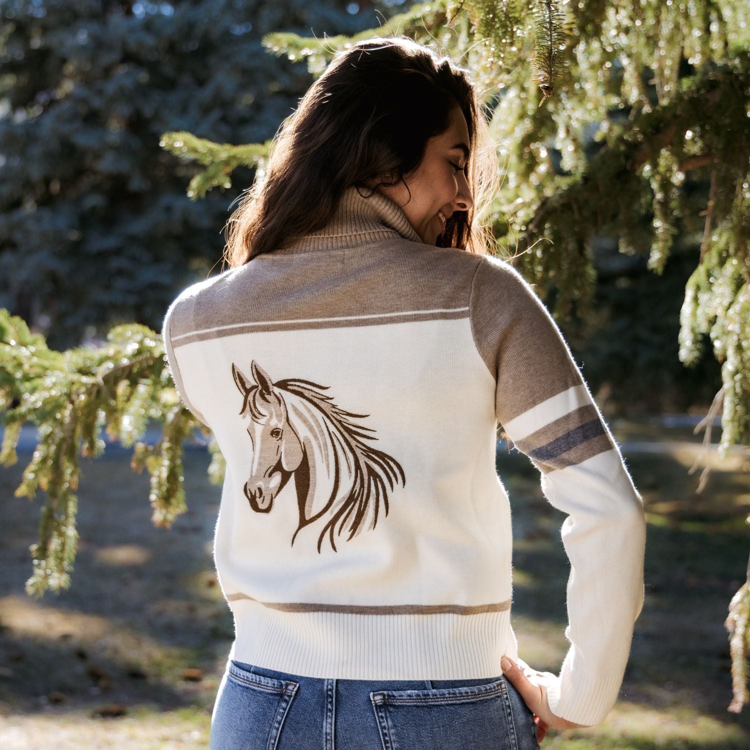 A woman viewed from behind wearing a cream and taupe knit sweater with a brown horse head graphic on the back, standing outdoors in front of evergreen trees.