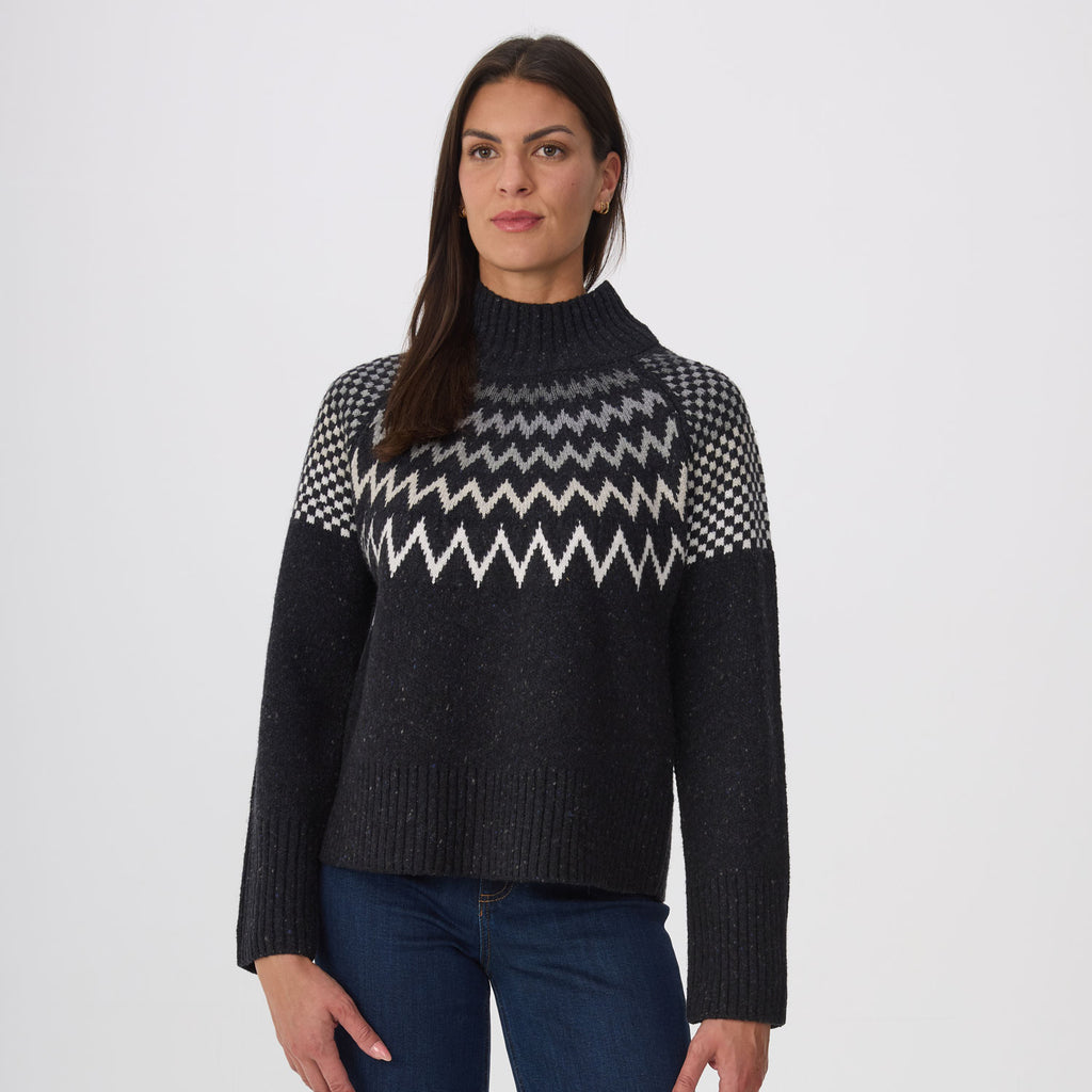 A woman modeling a heather black knit sweater with a white and black ombre geometric pattern across the chest and shoulders, paired with blue jeans, against a light gray studio background.
