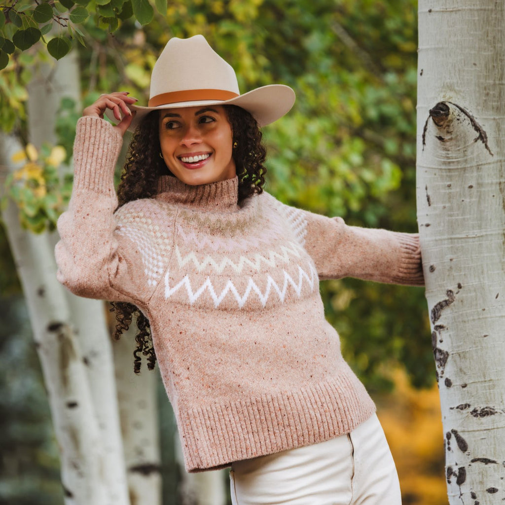 A woman outdoors wearing a chunky knit turtleneck sweater in a rose color with a white geometric zigzag pattern across the chest, paired with a beige cowboy hat and white pants, posing next to a aspen tree.