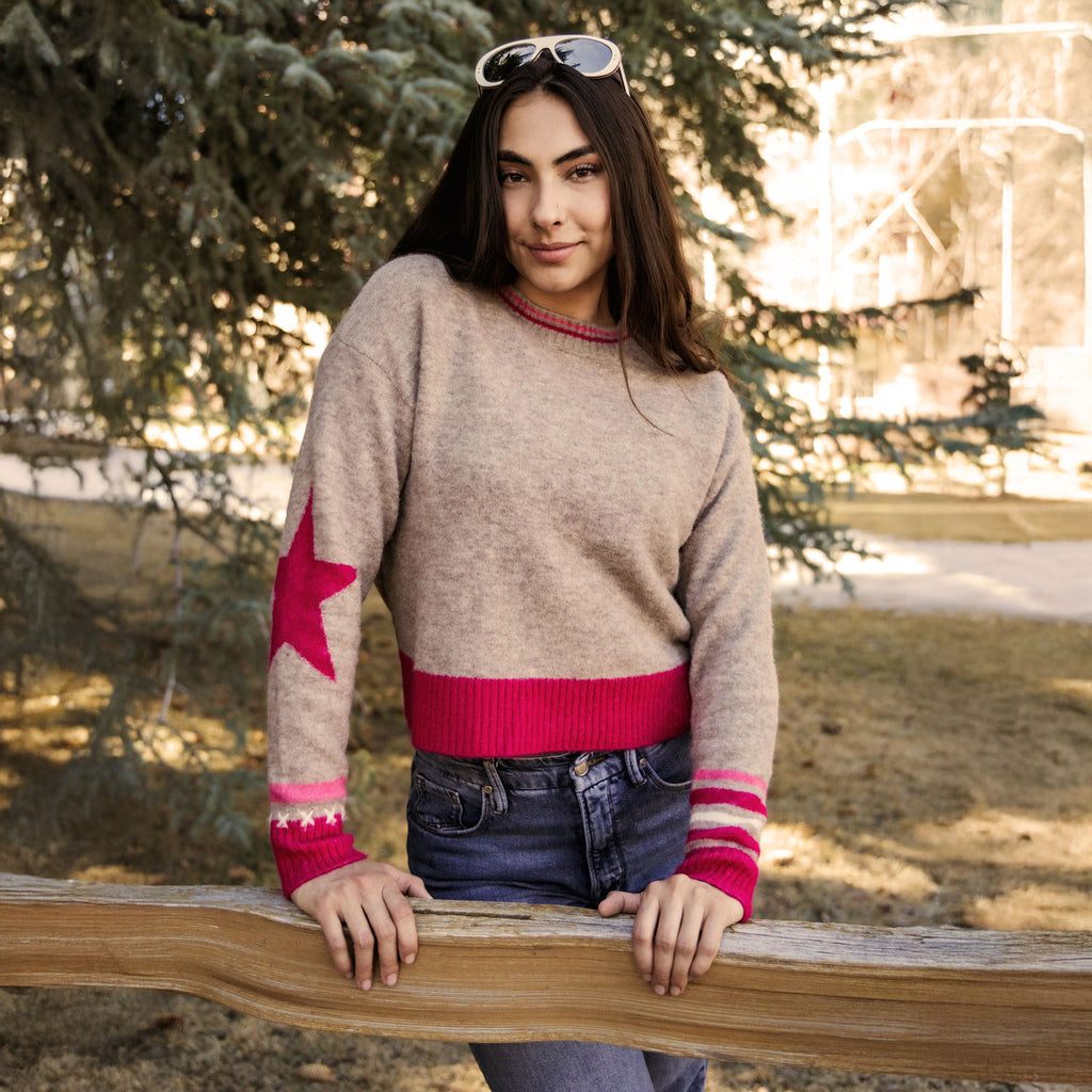 A woman wearing a beige knit sweater with a bright pink star on one sleeve, a pink ribbed hem and cuffs, and pink-and-white striped cuffs, standing outdoors behind a wooden fence.