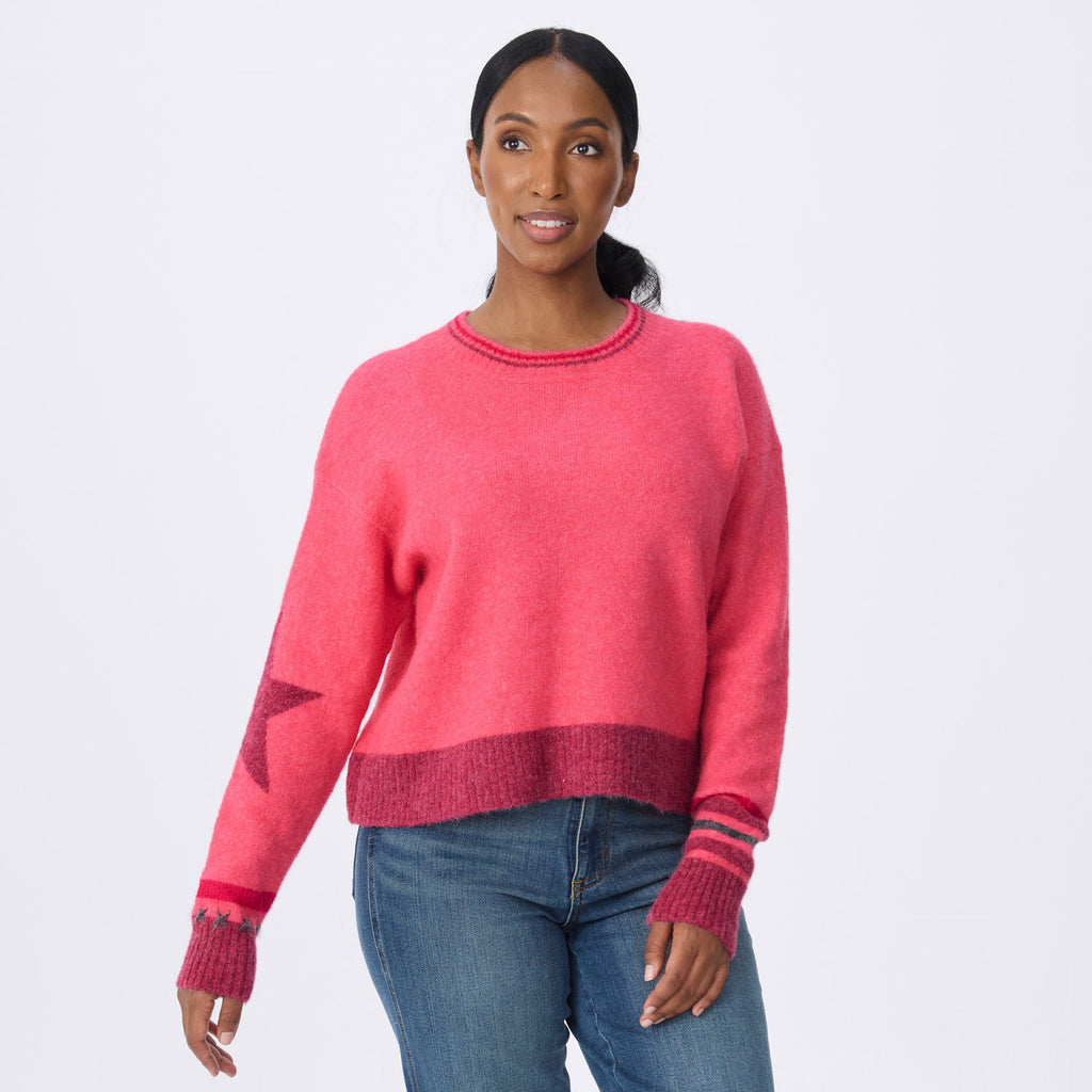 A woman modeling a bright pink crewneck sweater with darker pink ribbed trim, a star detail on the sleeve, and striped cuffs, paired with blue jeans against a light gray background.