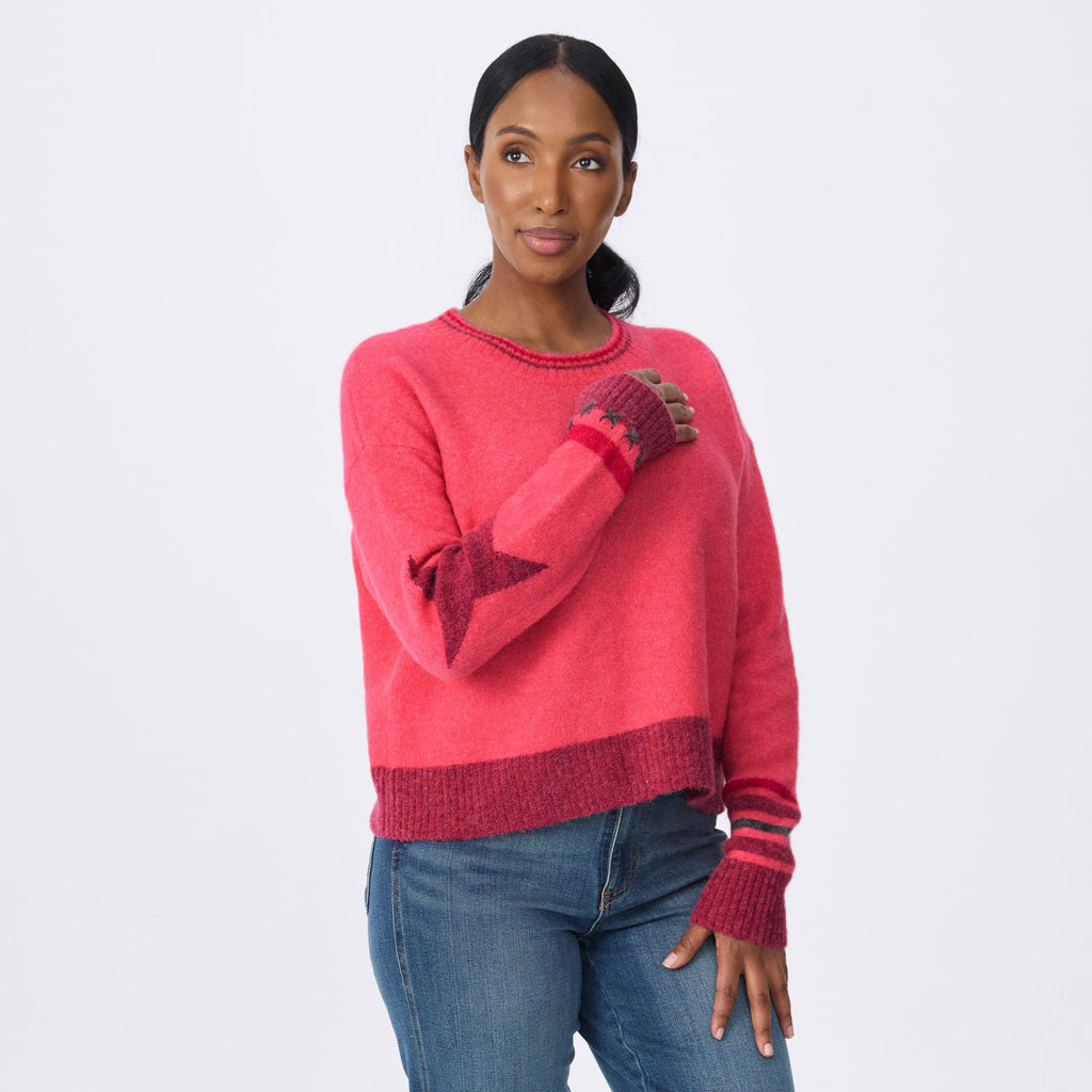 A woman modeling a pink knit sweater with geometric star patterns on the sleeves and striped cuffs, paired with blue jeans, against a plain light gray background.