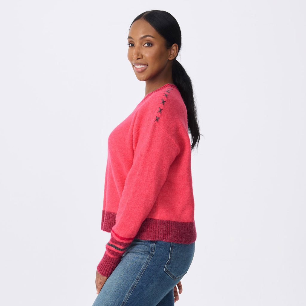 A woman wearing a bright pink knit sweater with dark pink ribbed trim and small dark star details on the shoulder, paired with blue jeans, photographed against a light gray studio background.