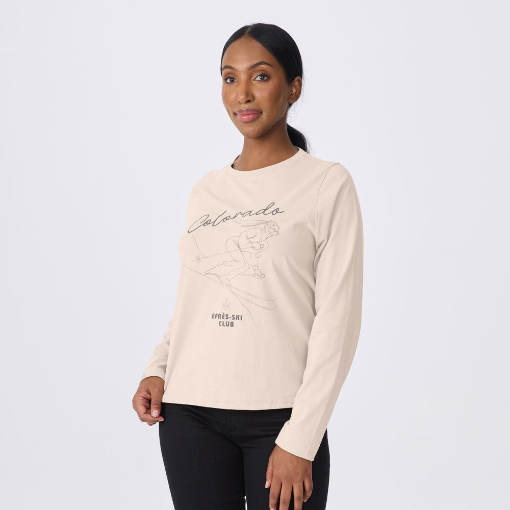 A woman modeling a light beige long-sleeve t-shirt with a black line art graphic of a skier and the text 'Colorado Alpine Ski Club' on the front, paired with black pants against a light gray studio background.
