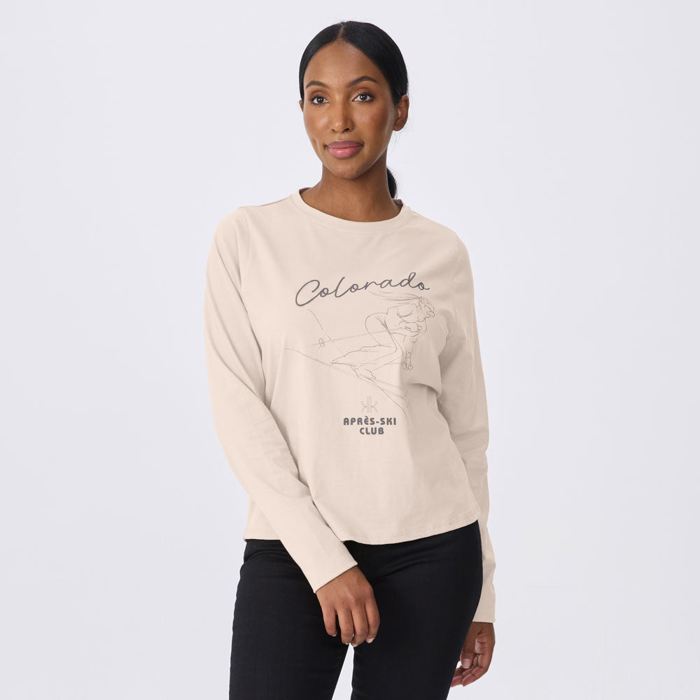 A woman modeling a cream-colored long-sleeve crewneck sweatshirt with a black graphic print that reads 'Colorado April Ski Club' on the front, paired with black pants, against a light gray studio background.