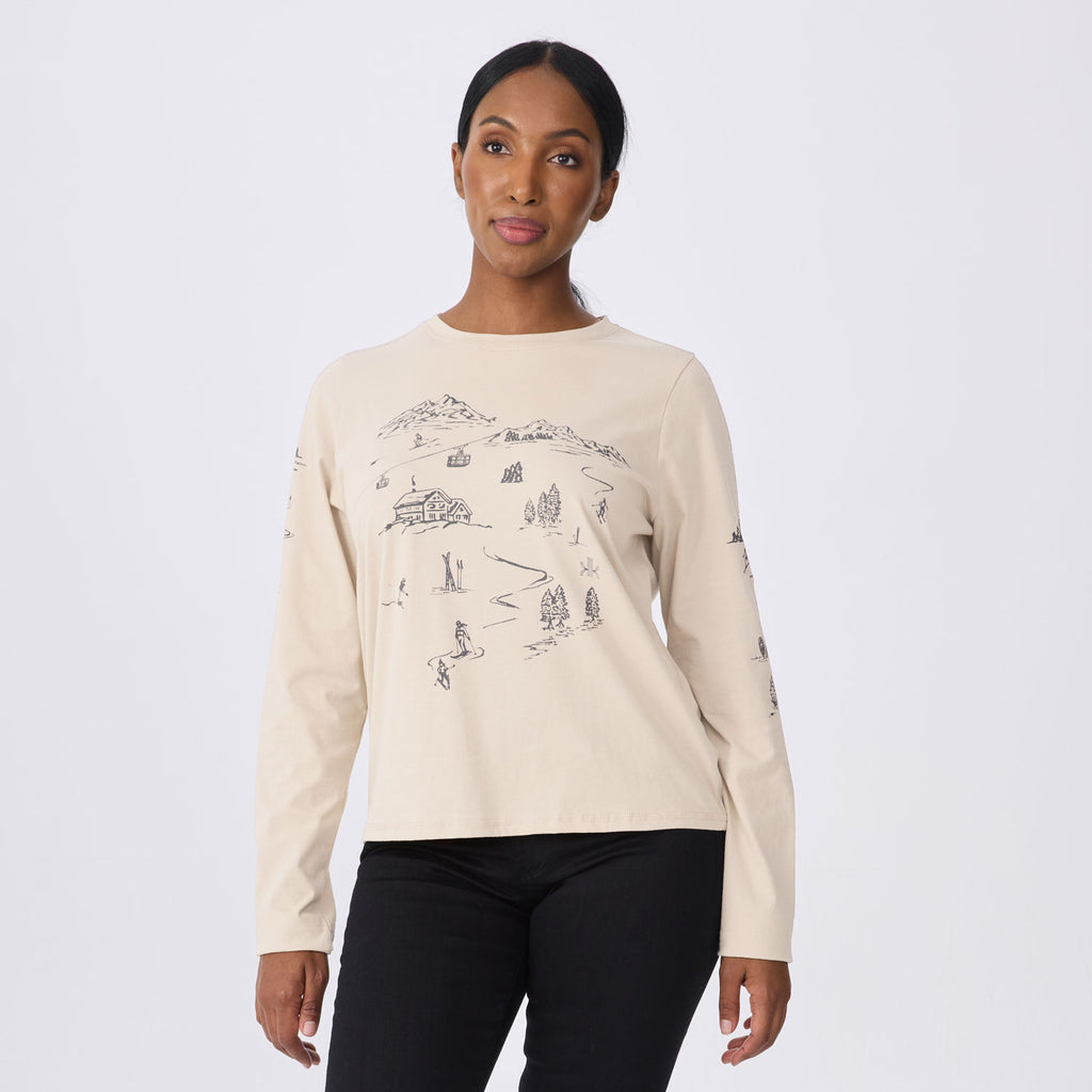 A woman modeling a cream-colored long-sleeve crewneck sweatshirt with a black line art graphic of a mountain cabin and skiers on the front, paired with black pants against a light gray studio background.