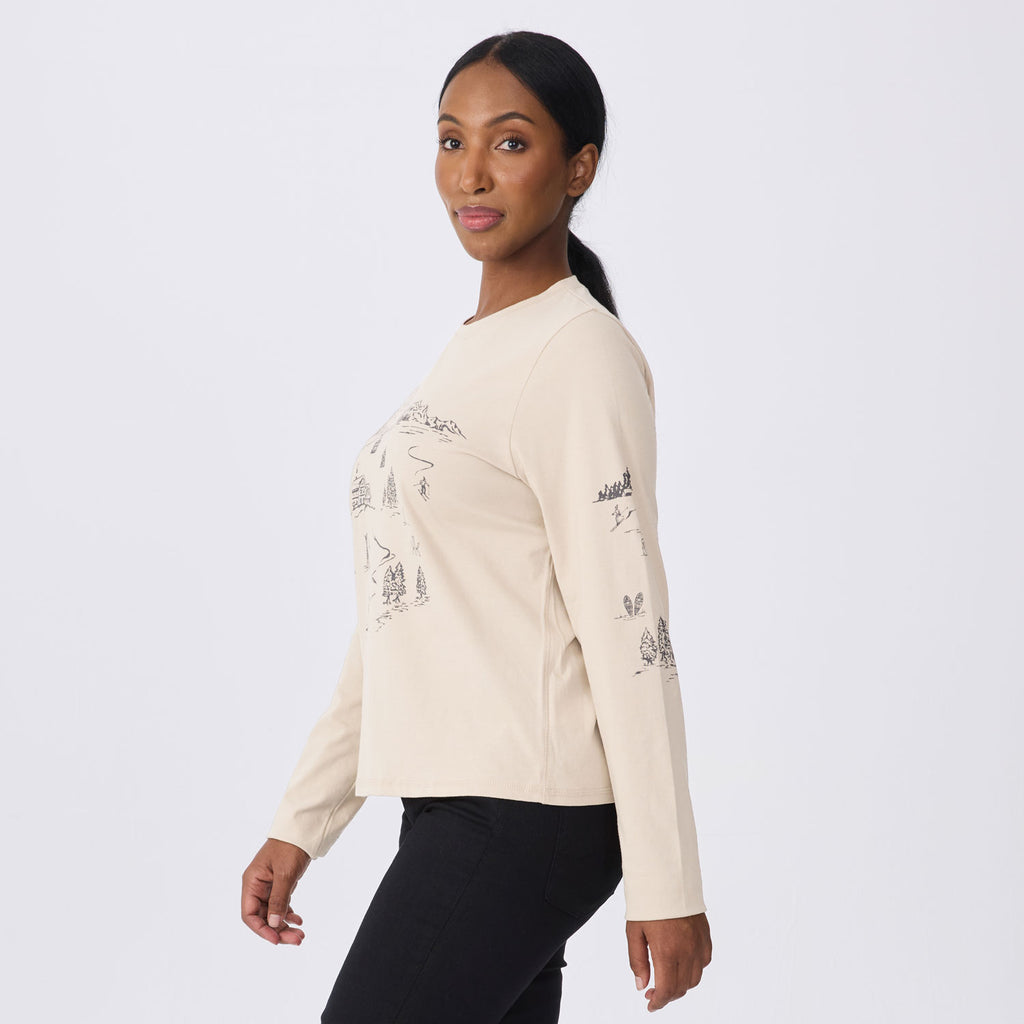 A woman wearing a cream-colored long-sleeve crewneck sweatshirt with black line-art illustrations of trees and mountains on the front and sleeves, paired with black pants, photographed against a light gray studio background.
