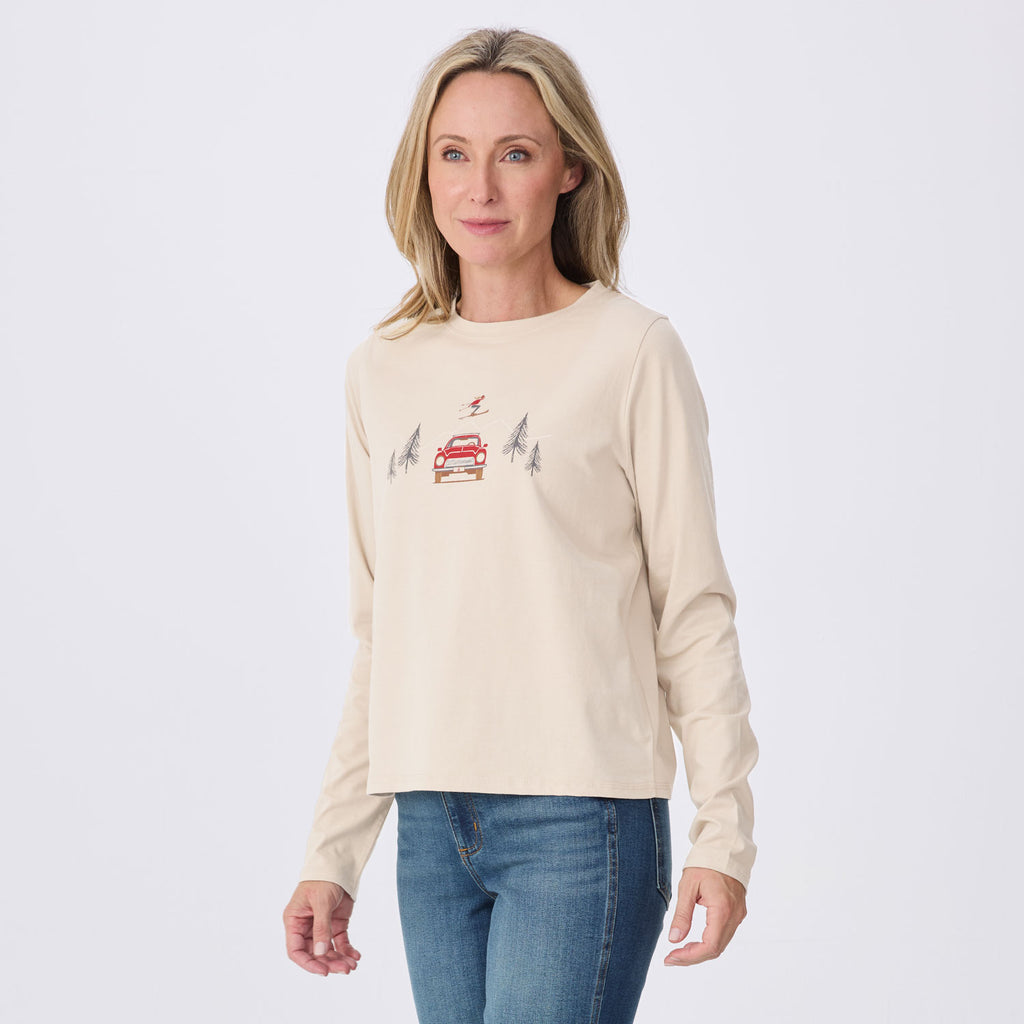 A woman modeling a cream-colored long-sleeve crewneck shirt with a small graphic of a red car and trees on the chest, paired with blue jeans against a light gray studio background.