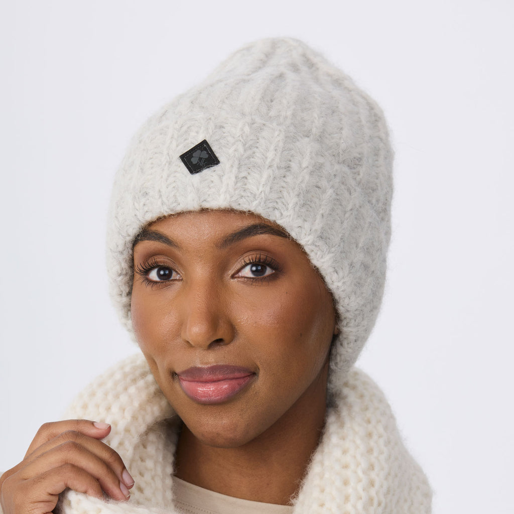A close-up of a woman wearing a chunky knit beanie in a light snow color, paired with a matching snow knit scarf. The beanie features a small black square logo patch on the front.
