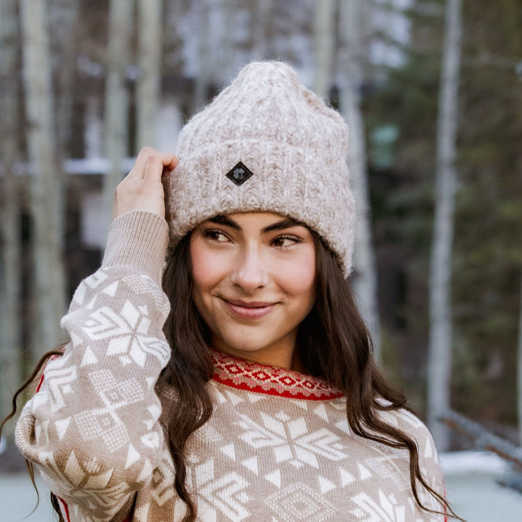 A woman wearing a beige knit beanie with a small black diamond logo and a beige patterned sweater with a red Nordic collar, posing outdoors in a snowy forest setting.