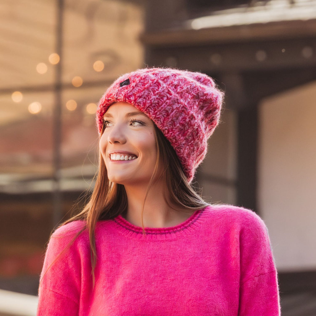 A smiling woman wearing a bright pink chunky knit beanie and a matching pink sweater, photographed outdoors with a softly blurred urban background.