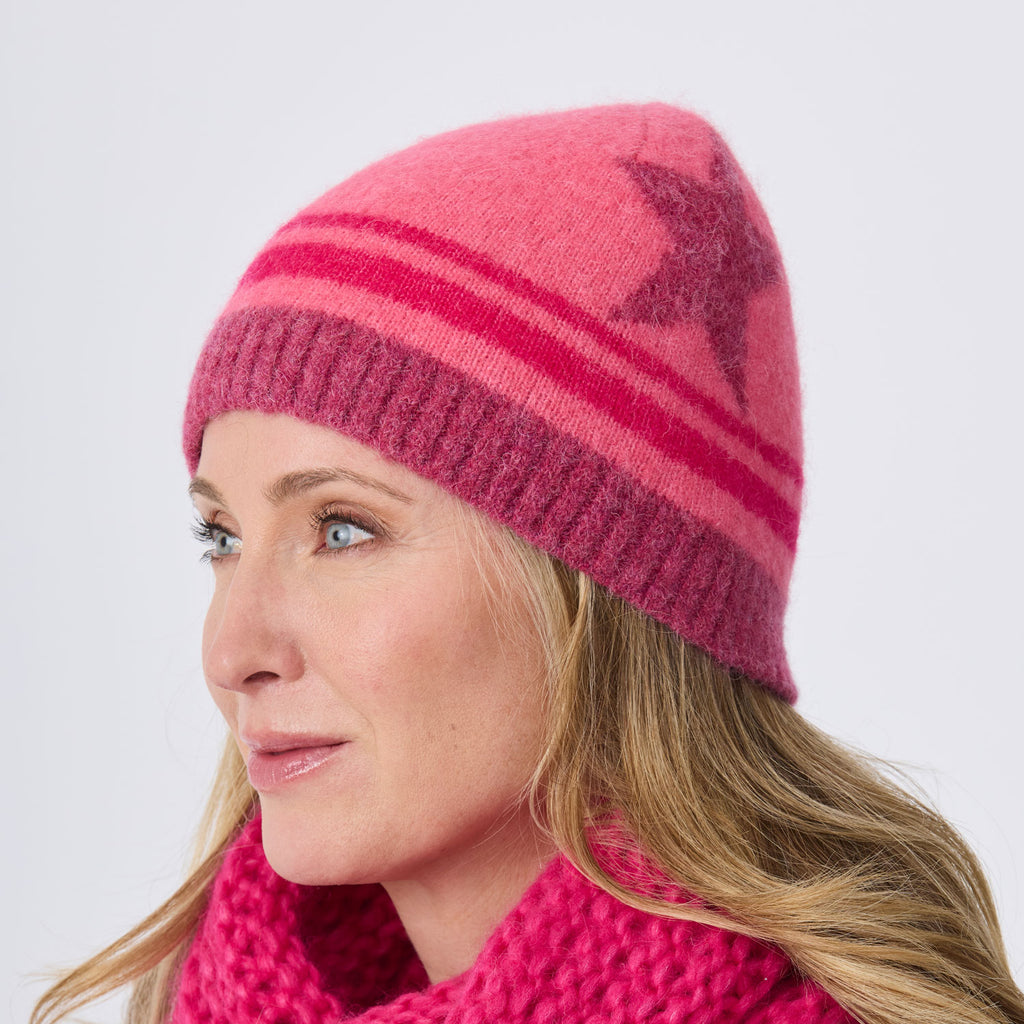 A woman wearing a pink knit beanie with darker pink stripes and a star motif, paired with a chunky pink scarf, shown in profile against a light gray background.