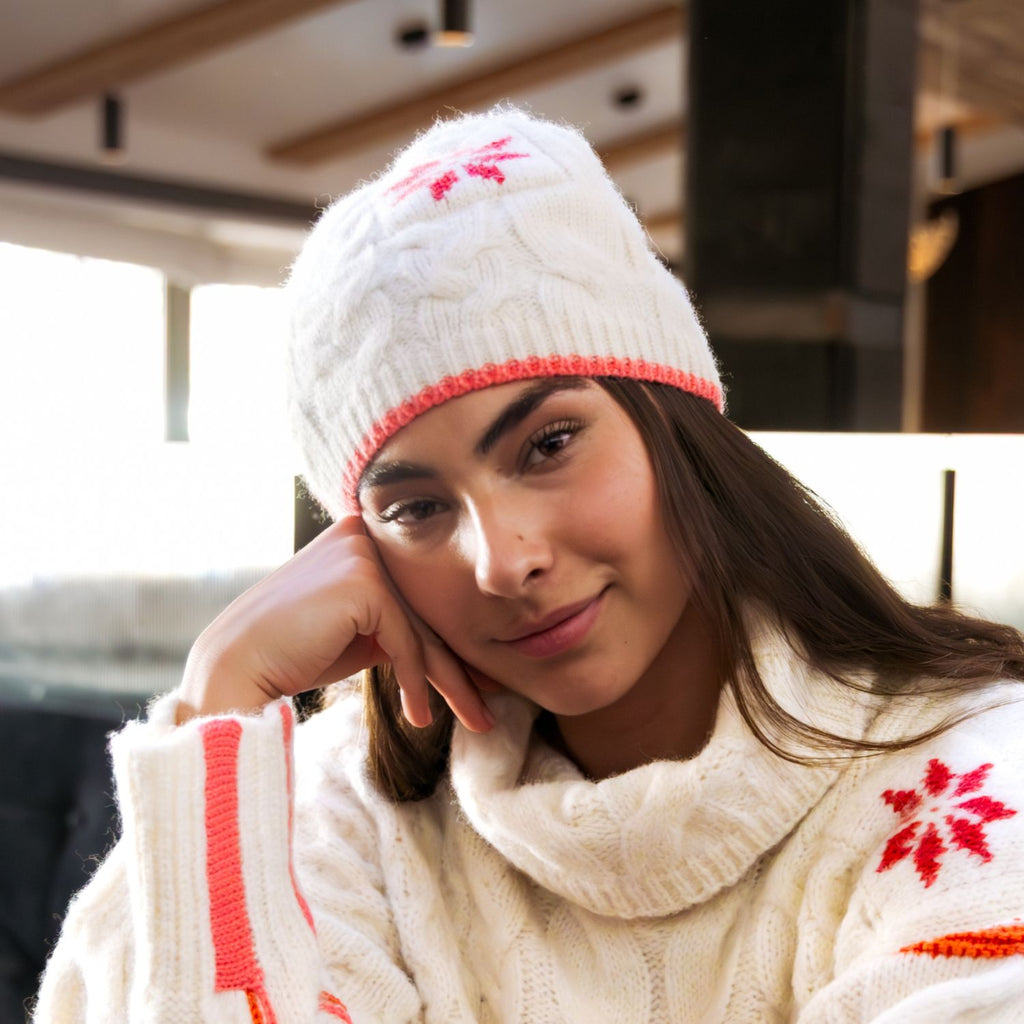 A woman wearing a cream cable-knit beanie with red snowflake motifs and a matching cream knit sweater, resting her head on her hand in a warm indoor setting.