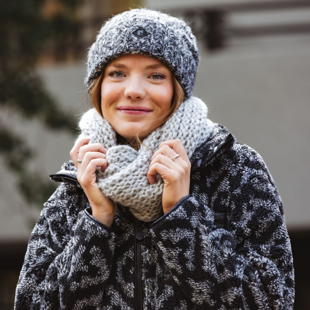 A woman wearing a thick light grey knit scarf, a chunky knit beanie, and patterned black-and-white jacket, holding the scarf in front of her face.