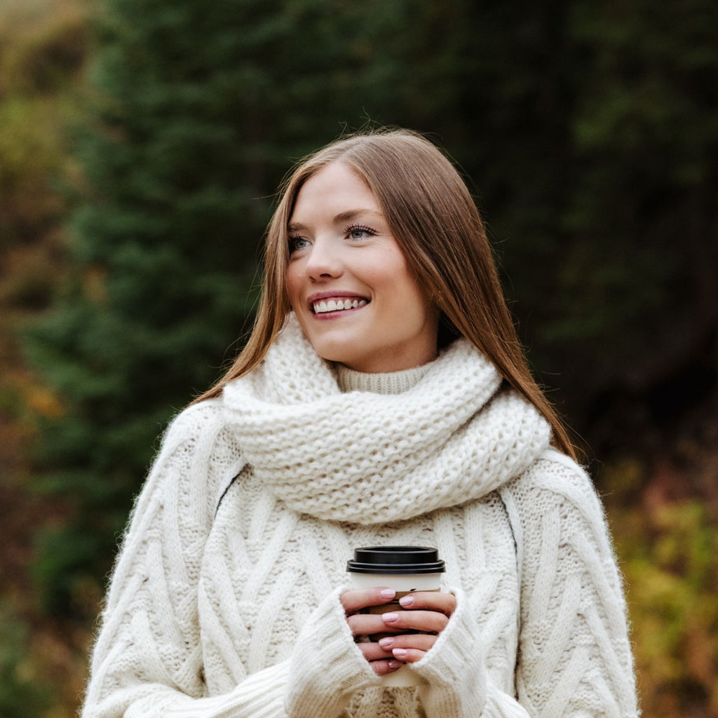 A smiling woman outdoors wearing a chunky cream knit scarf and matching sweater, holding a coffee cup against a blurred forest background.
