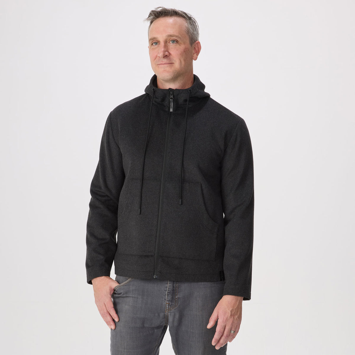 A man wearing a black zip-up hoodie with a high collar and drawstrings, paired with gray jeans, standing against a plain light gray studio background.