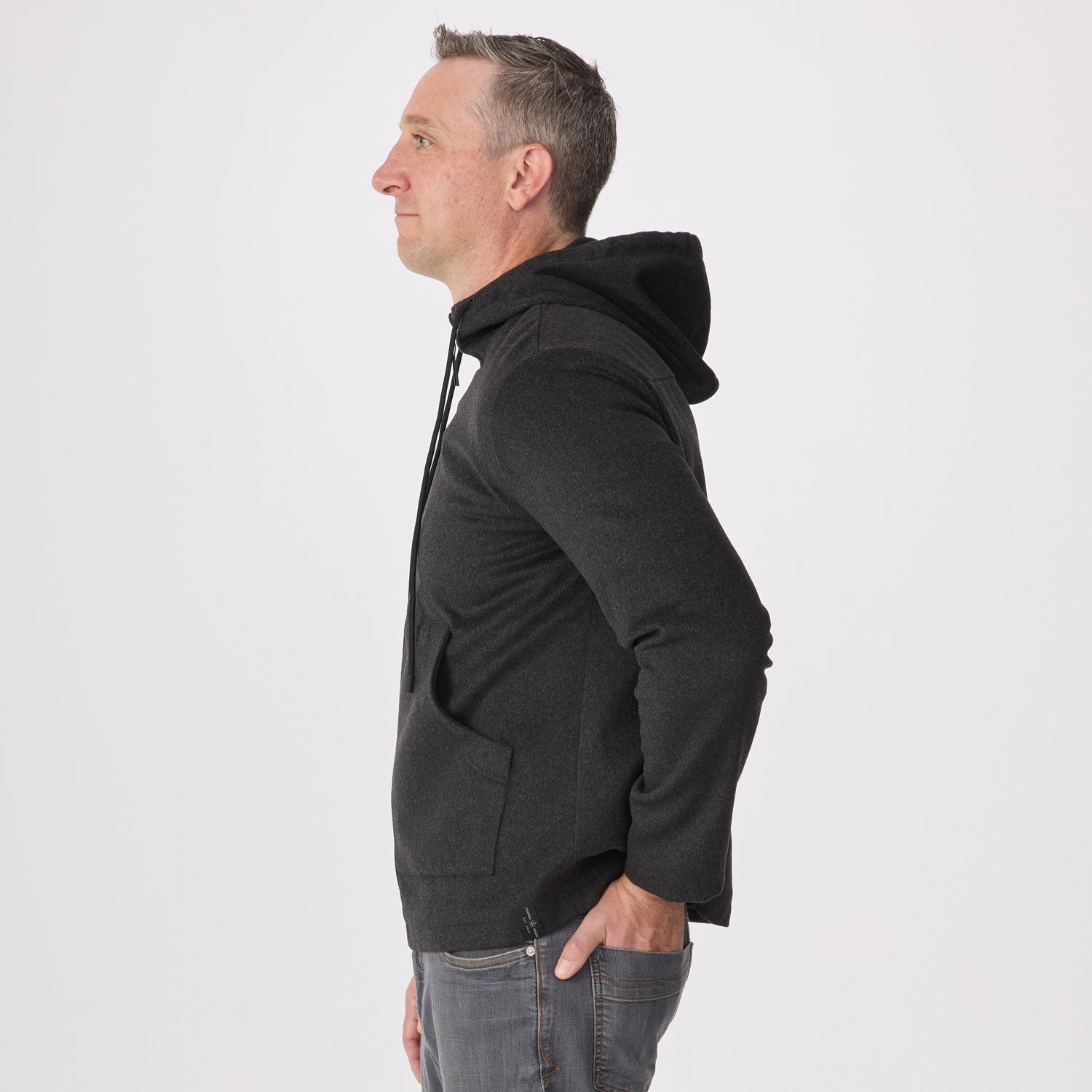 A man shown in profile wearing a dark charcoal gray hoodie and gray jeans, standing against a plain light gray studio background.