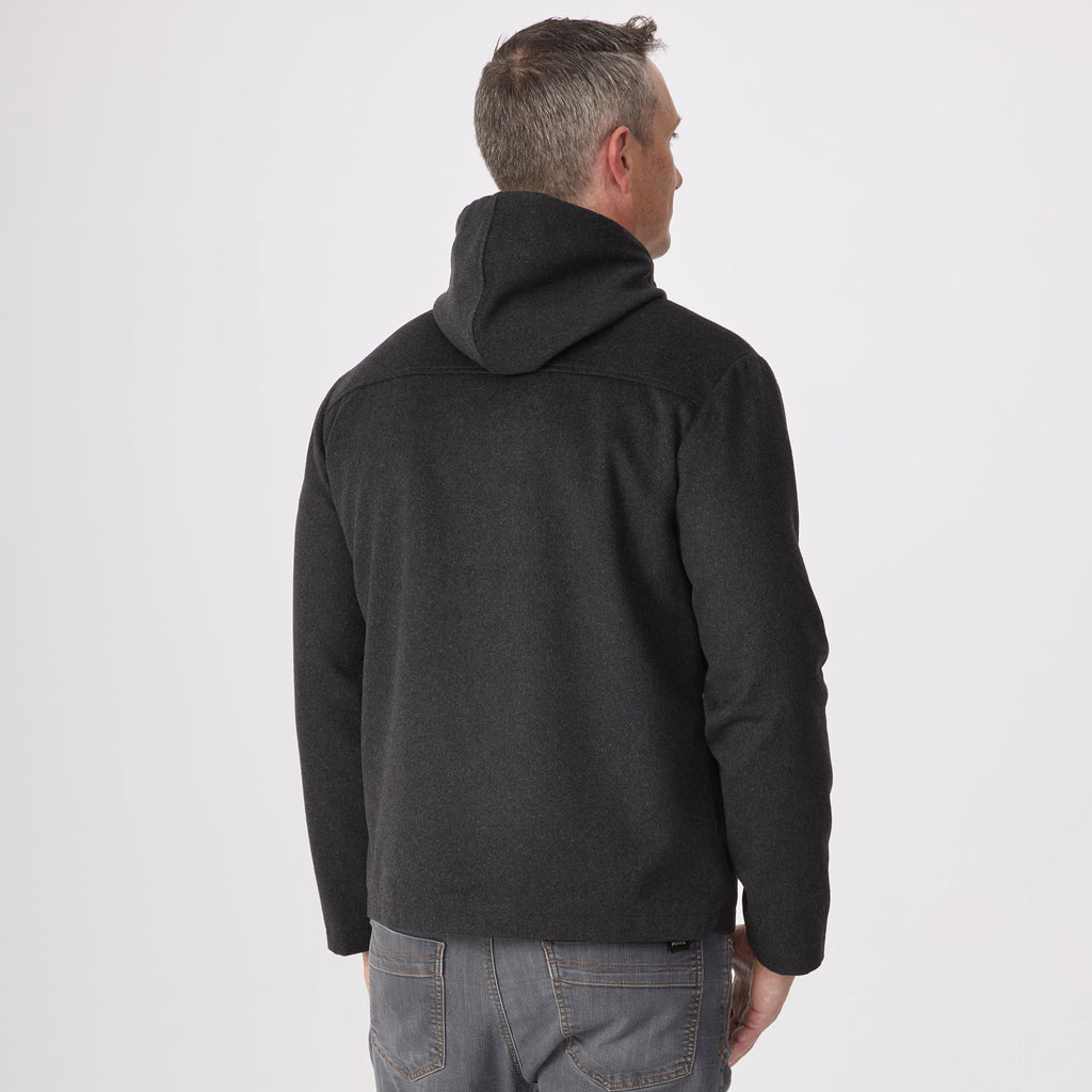 A man viewed from behind wearing a dark charcoal gray hoodie and gray jeans, standing against a plain light gray studio background.