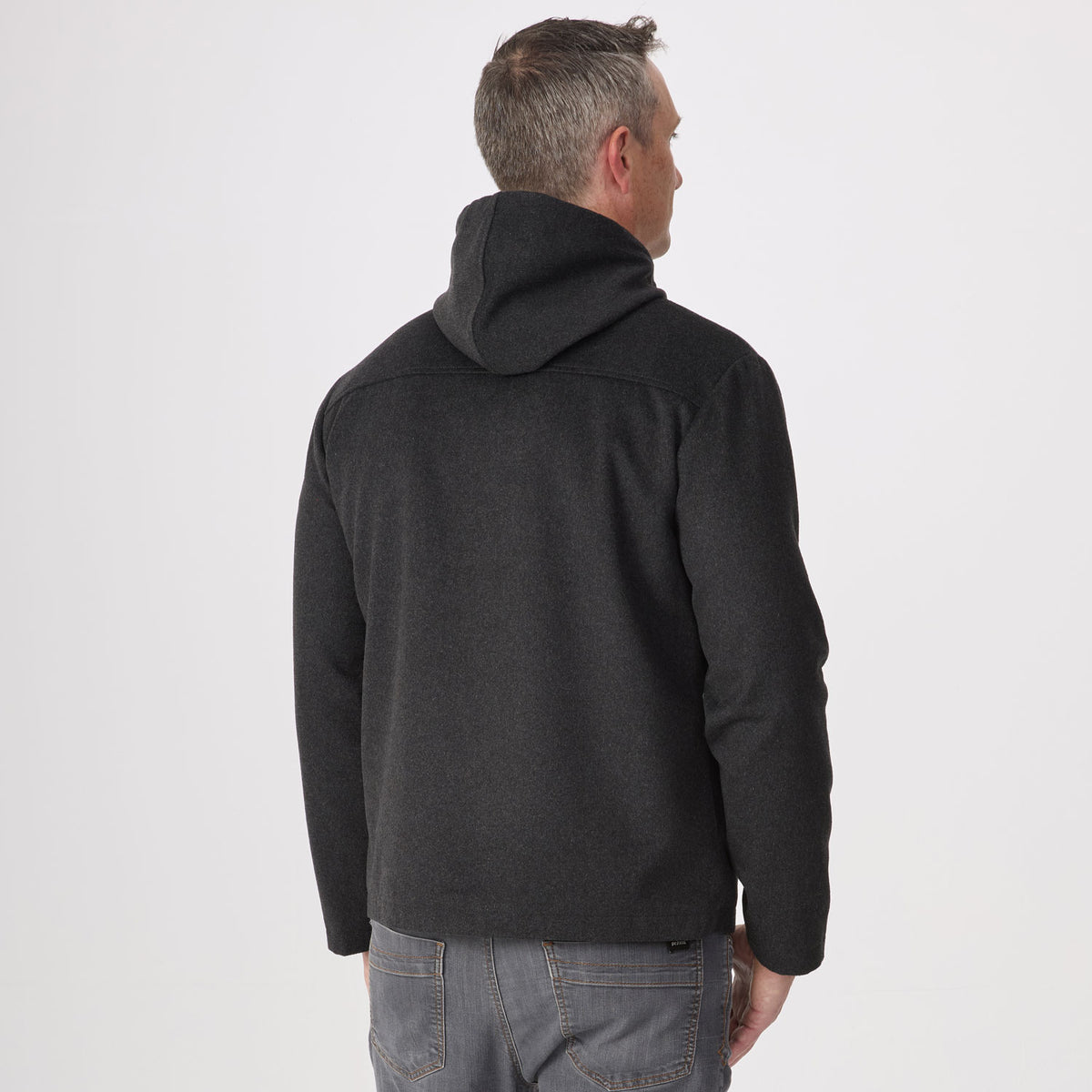 A man viewed from behind wearing a dark charcoal gray hoodie and gray jeans, standing against a plain light gray studio background.