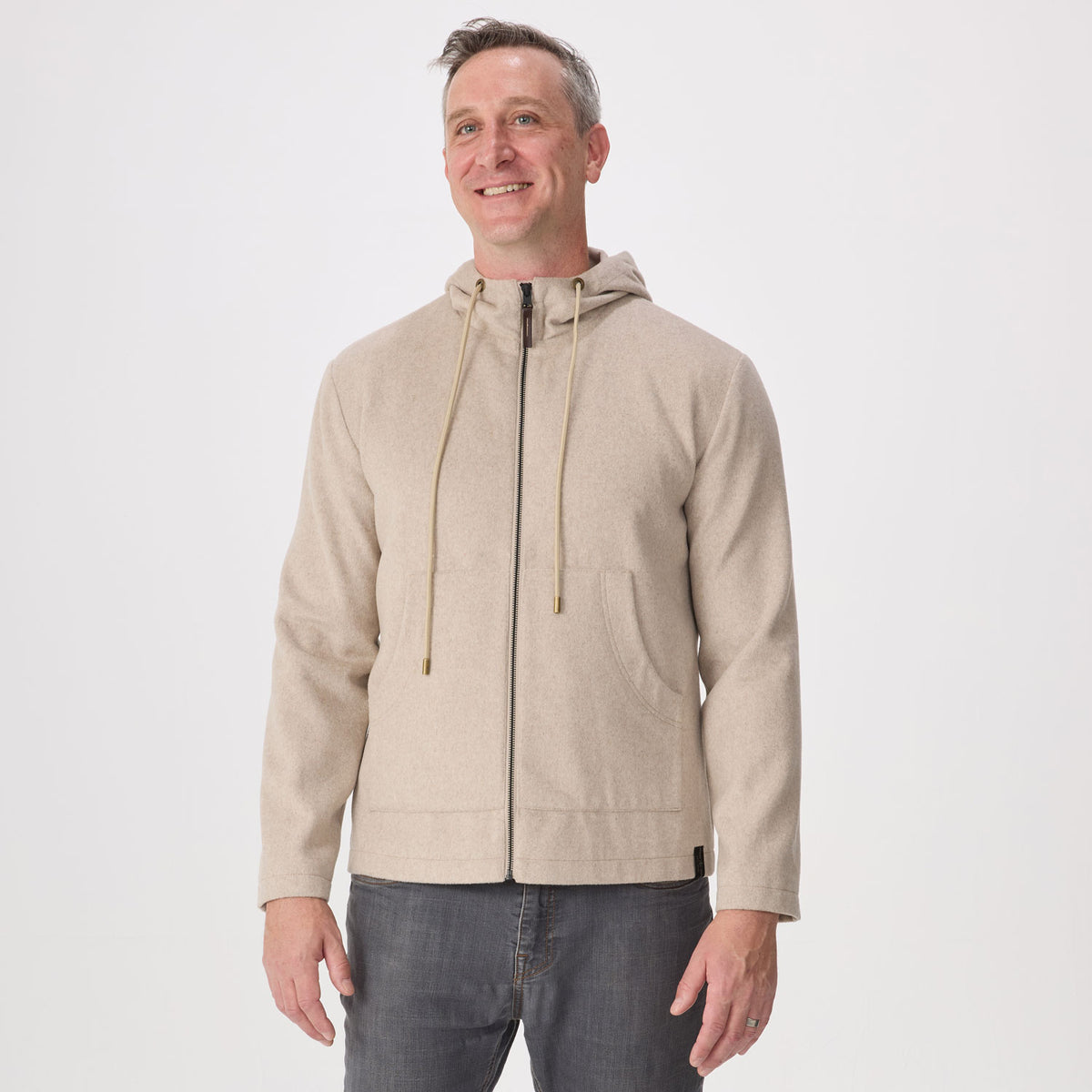 A man wearing a light beige zip-up hoodie with drawstrings and a kangaroo pocket, paired with dark gray jeans, standing against a plain white studio background.