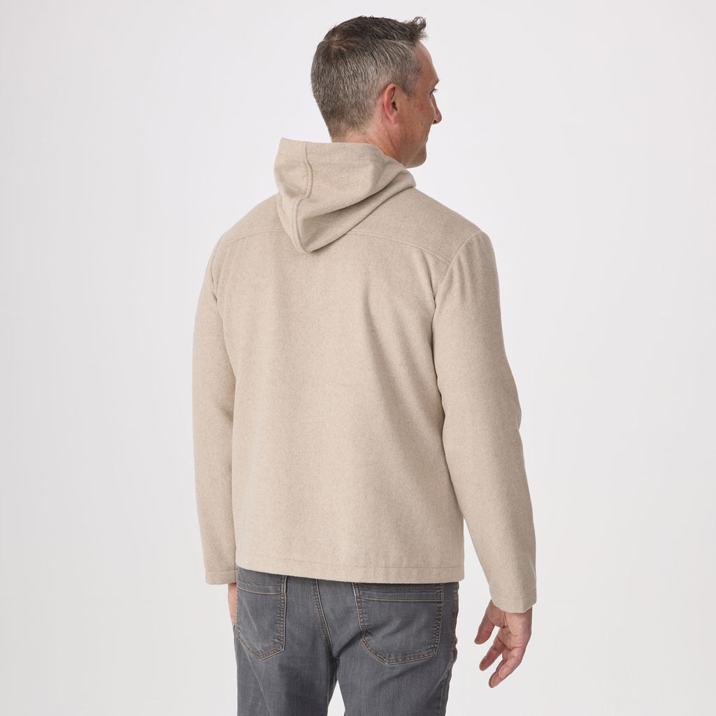 A man viewed from the back wearing a light beige hoodie and gray jeans, standing against a plain light gray studio background.