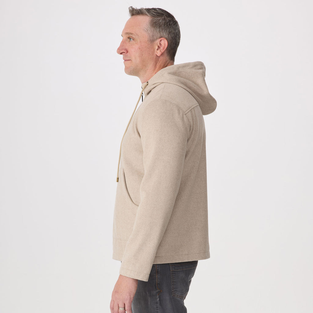 A man shown in profile wearing a light beige hoodie and dark jeans against a plain white studio background.