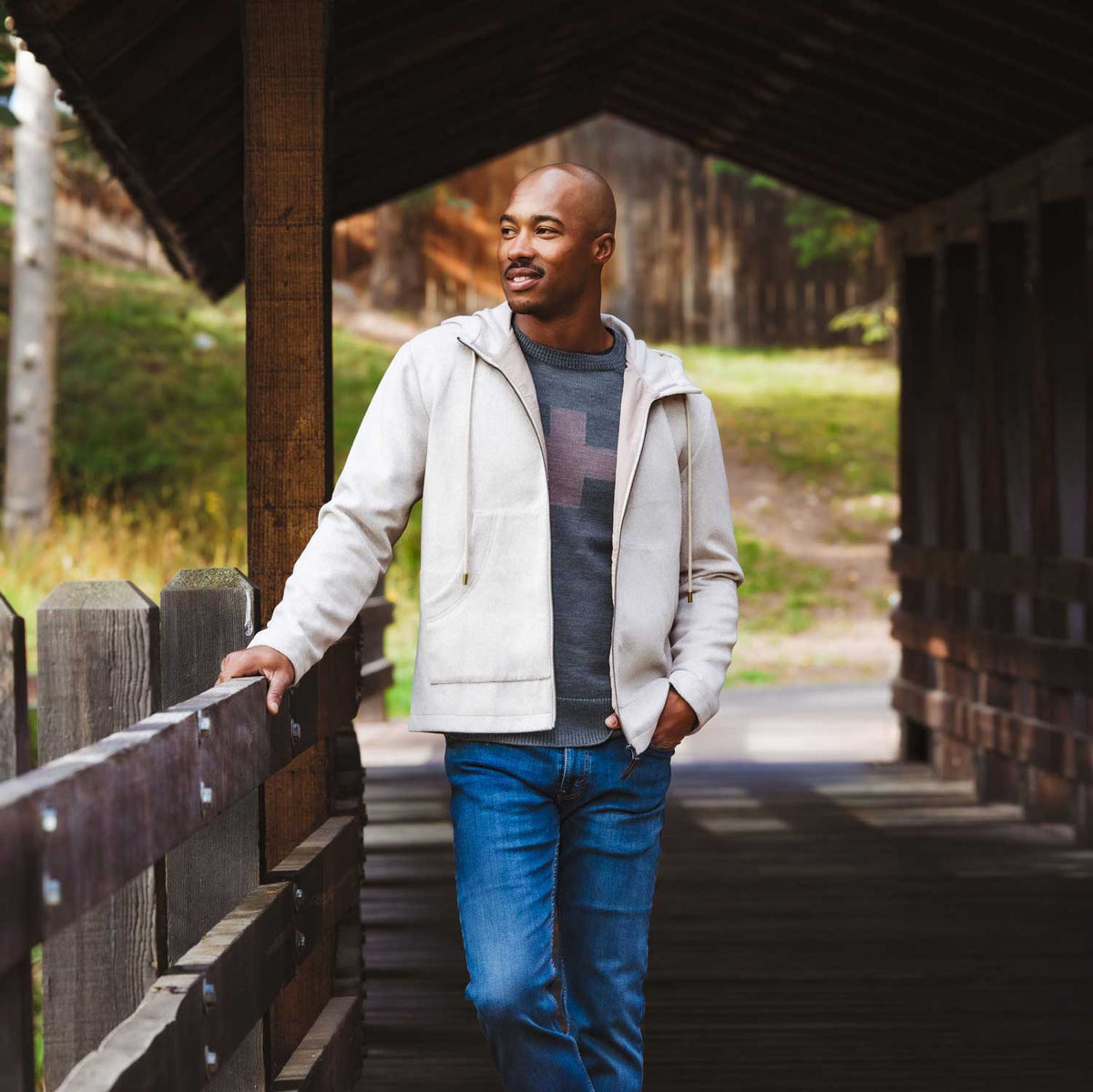 A man wearing a light beige zip-up hoodie over a gray sweater and blue jeans, standing on a wooden covered bridge.