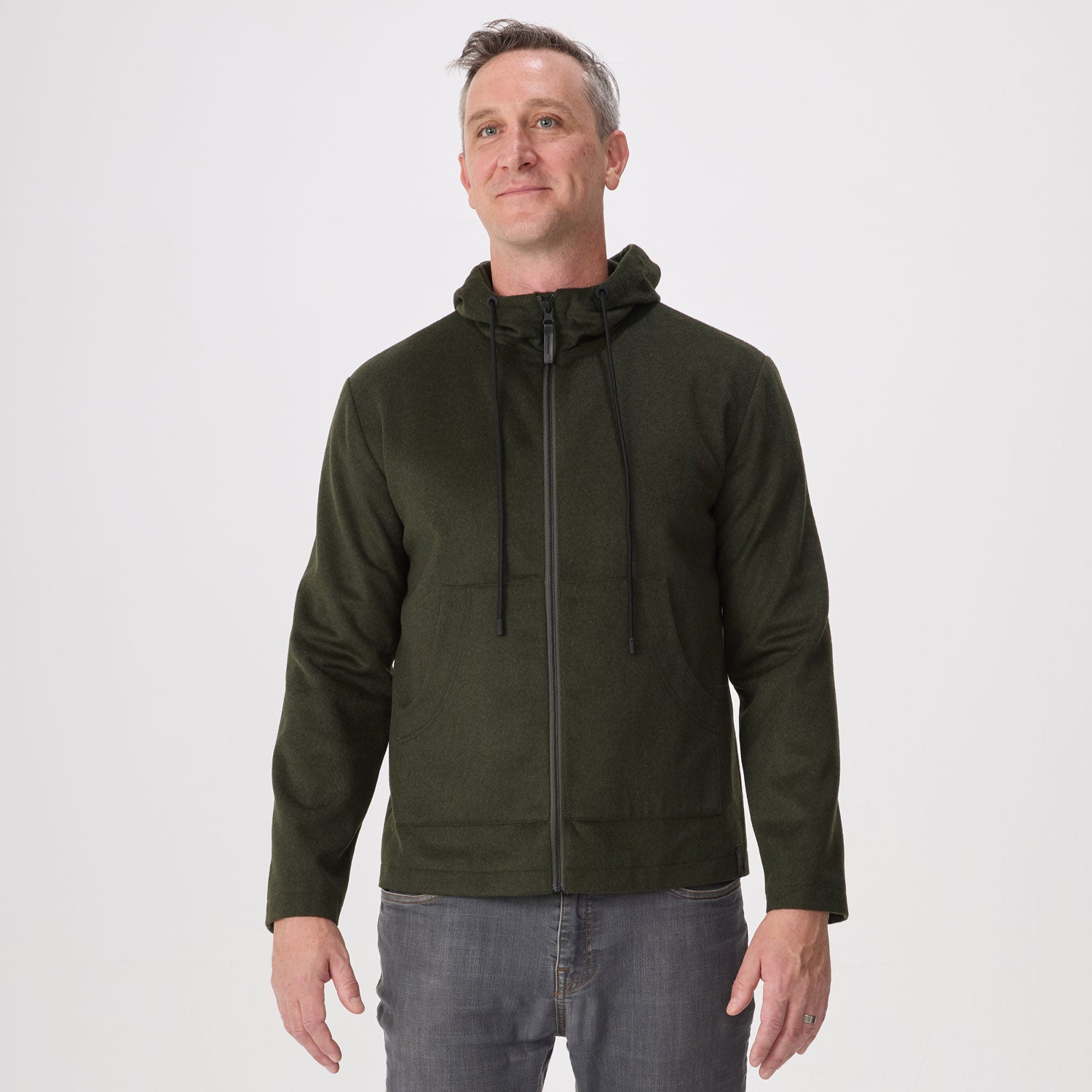 A man wearing an olive green zip-up hoodie with black drawstrings, paired with gray jeans, standing against a light gray studio background.