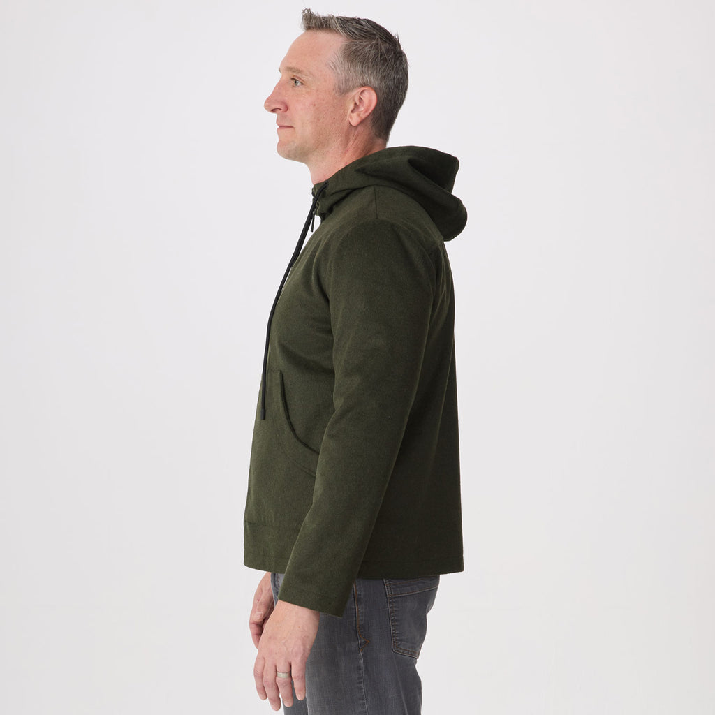 A man shown in profile wearing an olive green hoodie and gray jeans against a plain light gray studio background.