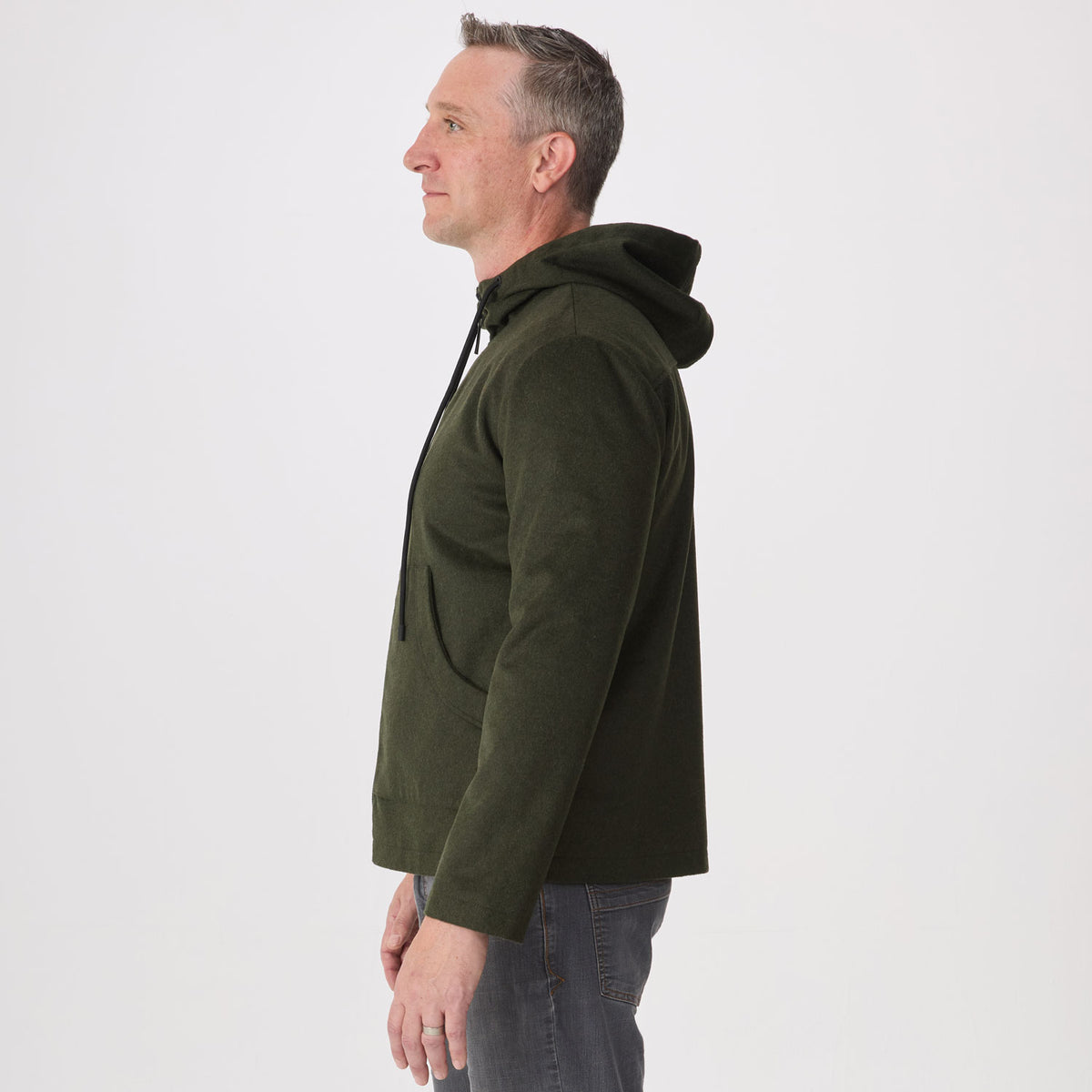 A man shown in profile wearing an olive green hoodie and gray jeans against a plain light gray studio background.
