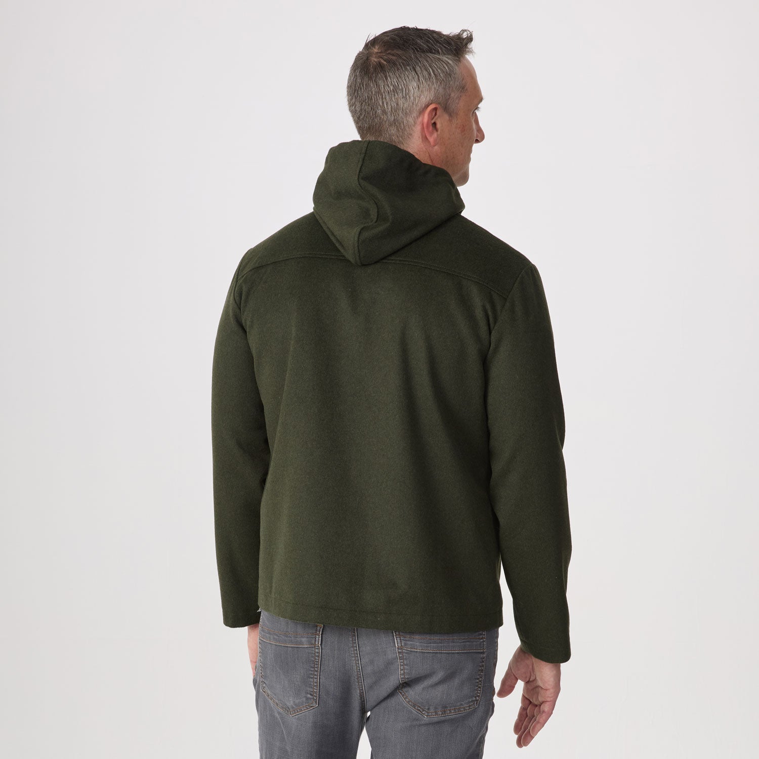 A man viewed from the back wearing an olive green hoodie and gray jeans, standing against a plain light gray studio background.