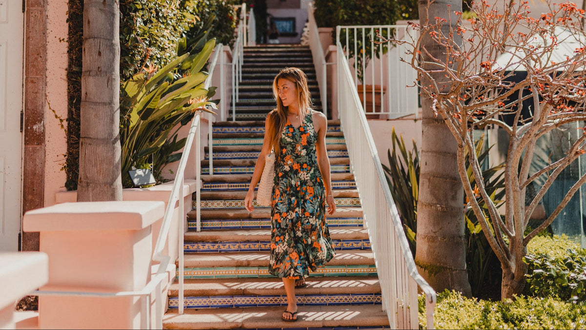 60% Off Labor Day Weekend Sale. A woman wearing a sleeveless floral maxi dress walks down a patterned outdoor staircase between palm trees and tropical plants.
